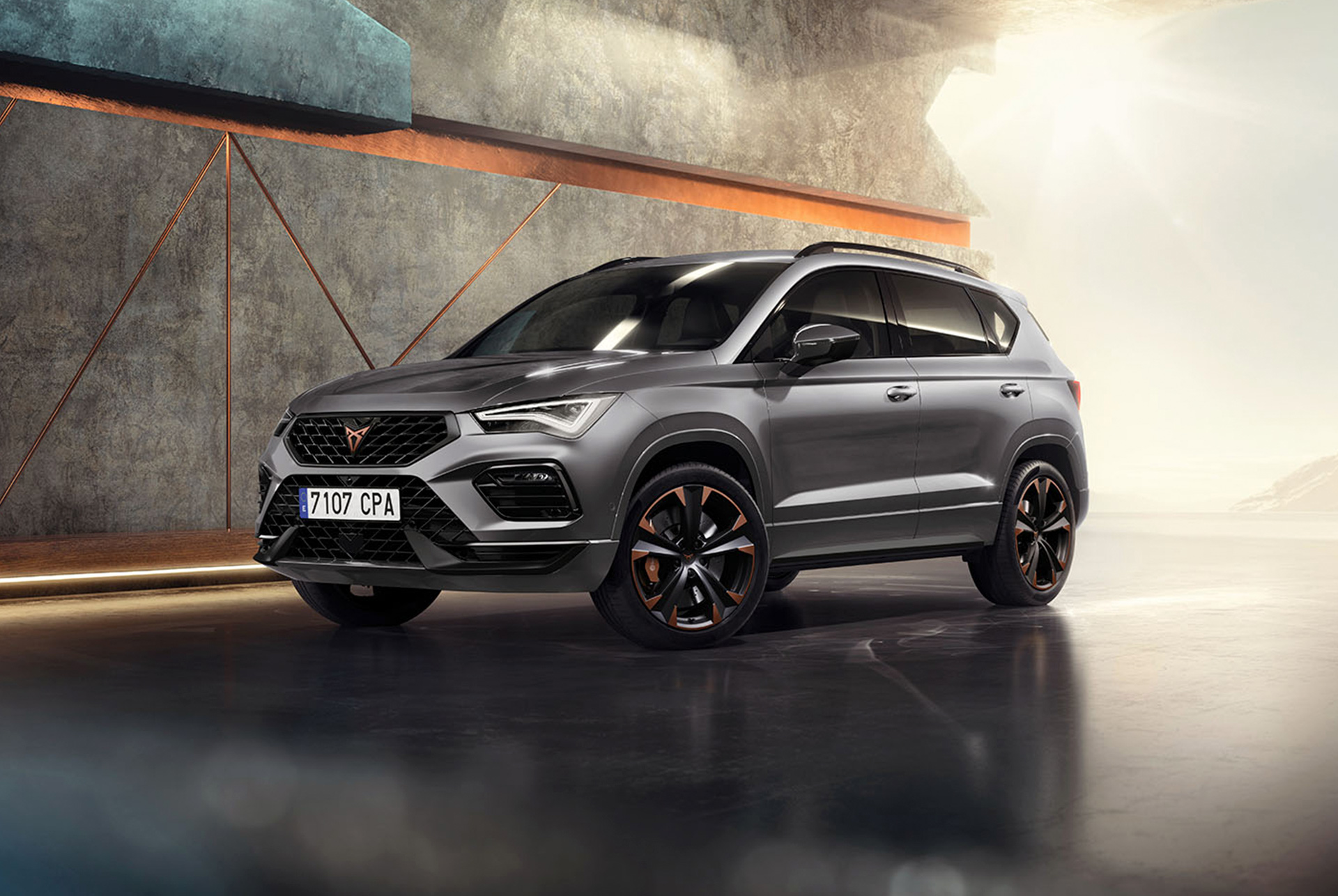 CUPRA Ateca Graphite Grey krāsā, sportisks un kompakts SUV modernā, industriālā vidē. Automobiļa dizainu raksturo aerodinamiskas līnijas, LED priekšējie lukturi un oriģināli vieglmetāla diski ar vara akcentiem. Fonā ir ģeometrisks sienas dizains, kas kontrastē ar automobiļa eleganto dizainu.